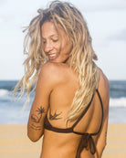 Eine Frau mit langen, gewellten blonden Haaren und sichtbaren Armtattoos lächelt an einem Sandstrand und trägt das Main Design Pasta Bikini Oberteil. Das Meer und der Himmel sind sanft im Hintergrund verschwommen.