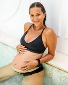 Eine schwangere Frau in einem schwarzen Pasta Bikini Oberteil von Main Design, hergestellt aus recycelten Materialien, lächelt in einem Pool mit nassen Haaren und goldenen Ohrringen, stützt eine Hand auf ihren Bauch und die andere auf den Rand und wirkt entspannt und glücklich.
