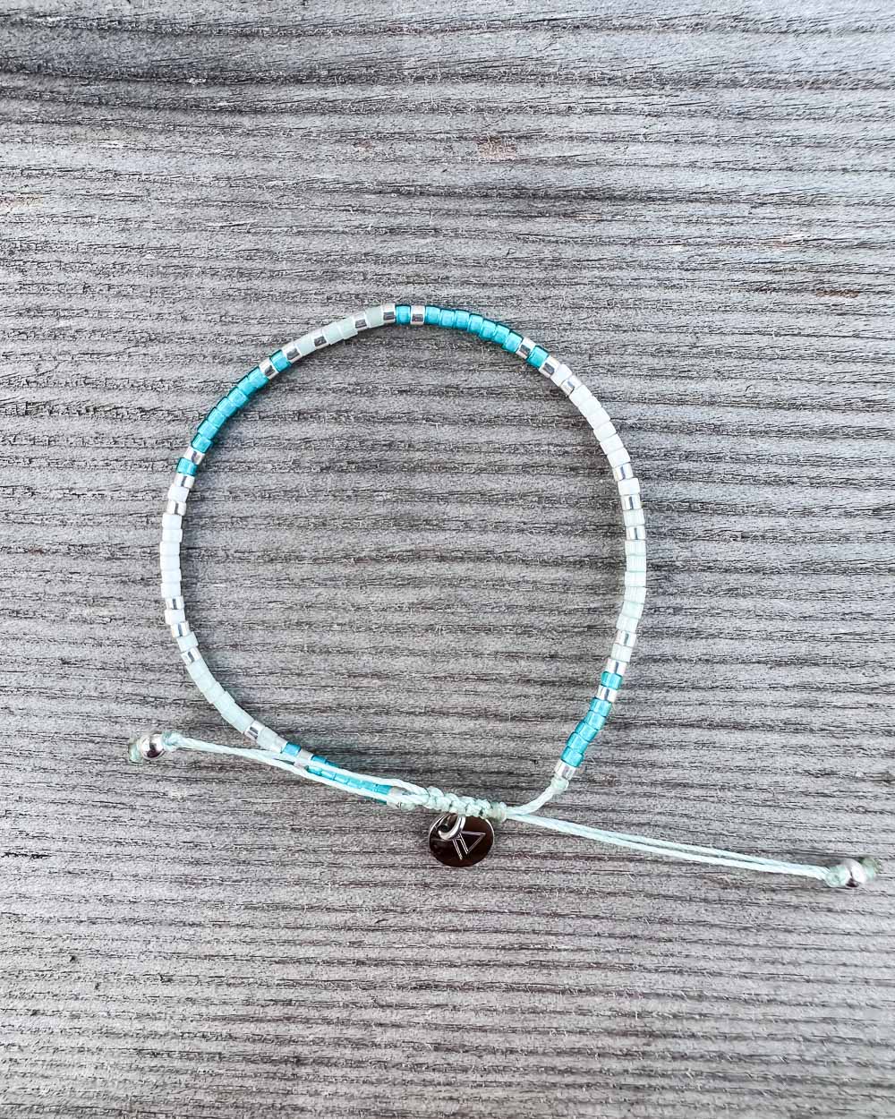 Das Mama und Mini Armband Wave von Main Design ist ein handgefertigtes, nachhaltiges Schmuckstück mit türkisfarbenen und silbernen Perlen sowie rundem Metallanhänger - perfekt präsentiert auf grauem Holz.