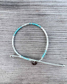 Das Mama und Mini Armband Wave von Main Design ist ein handgefertigtes, nachhaltiges Schmuckstück mit türkisfarbenen und silbernen Perlen sowie rundem Metallanhänger - perfekt präsentiert auf grauem Holz.