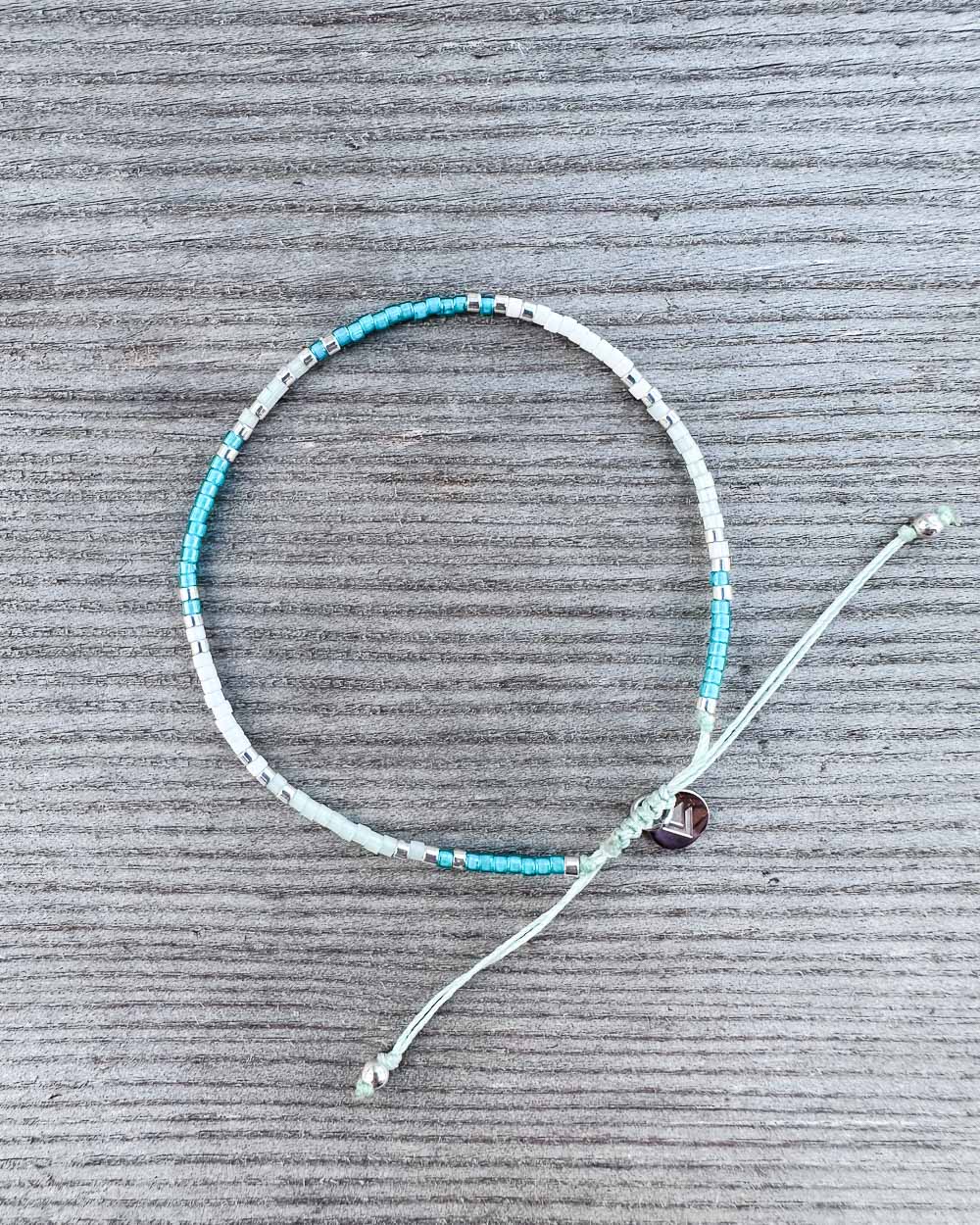 Das "Mama und Mini Armband Wave" von Main Design mit türkisfarbenen, silbernen und weißen Perlen sowie verstellbarem Verschluss wird auf grauer Oberfläche präsentiert - ideal als nachhaltiger Schmuck.