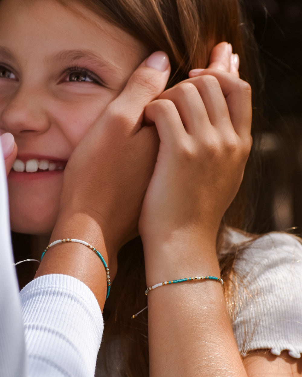 Ein lächelndes junges Mädchen mit langen braunen Haaren und einem weißen Oberteil berührt ihr Gesicht. Sie trägt das Mama und Mini Armband Wave von Main Design. Die Hand einer anderen Person berührt sanft ihre Wange.