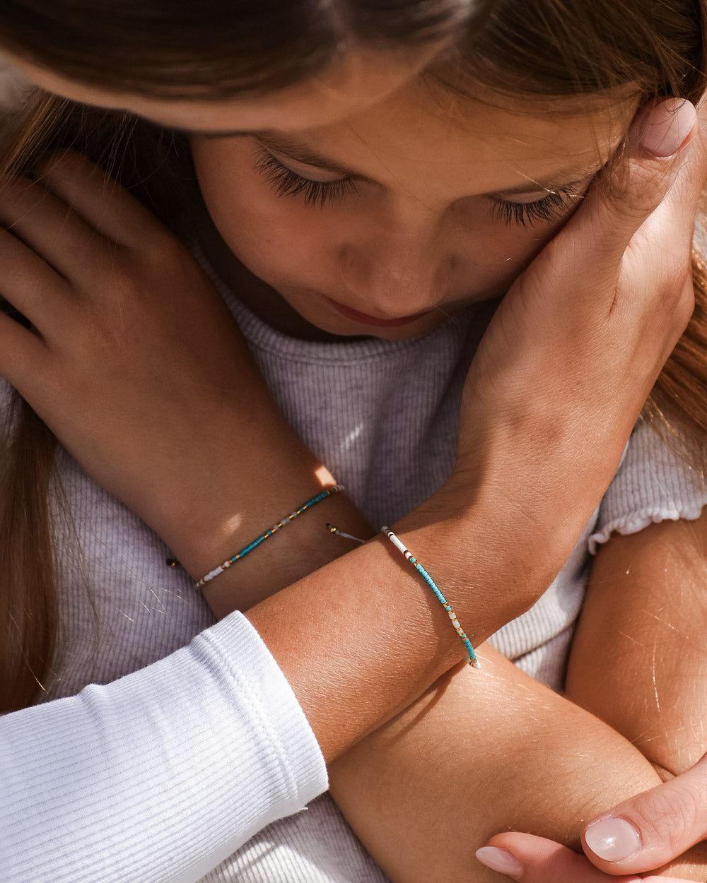 Ein kleines Mädchen mit geschlossenen Augen wird durch die Umarmung eines Erwachsenen getröstet. Beide tragen das bunte Mama und Mini Armband Wave von Main Design und fangen einen Moment der Wärme und Beruhigung mit nachhaltigem Schmuck ein.
