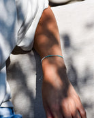 Eine Person trägt das filigrane Mama und Mini Armband Wave von Main Design in Türkis und legt ihren Arm auf hellen Stoff, während Sonnenlicht und Schatten sanft über die Haut spielen - ein Beispiel für nachhaltigen Schmuck.