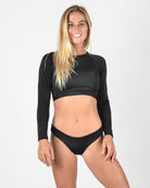 Eine Frau steht lächelnd in die Kamera. Sie hat langes blondes Haar und trägt ein schwarzes, langärmliges Lycra-Crop-Top von Maria Langarm mit einem schwarzen Bikinihöschen von Main Design. Der Hintergrund ist schlicht weiß.