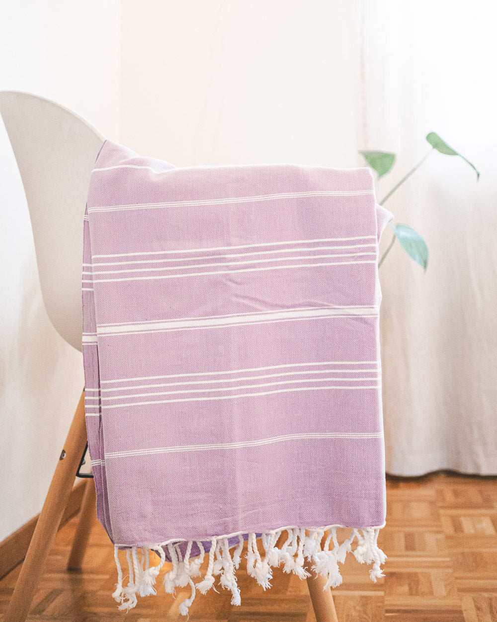 Das Maxi Strandtuch De La Mer von Main Design aus 100% Baumwolle mit weißen Fransen, OEKO-TEX zertifiziert, ist über einen weißen Stuhl auf Parkett drapiert. Im Hintergrund sind eine grüne Pflanze und ein weißer Vorhang sichtbar.