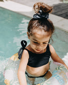 Ein junges Mädchen mit Haarknoten trägt einen Mia Mädchen Bikini von Main Design und benutzt einen bunten aufblasbaren Schlauch, während sie im seichten Poolwasser sitzt. Sie blickt nach unten, während das Sonnenlicht einen sanften Schein auf die Szene wirft.