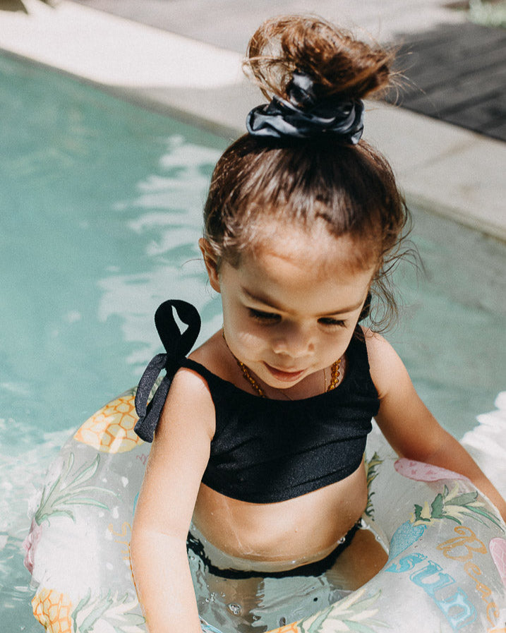 Ein junges Mädchen mit Haarknoten trägt einen Mia Mädchen Bikini von Main Design und benutzt einen bunten aufblasbaren Schlauch, während sie im seichten Poolwasser sitzt. Sie blickt nach unten, während das Sonnenlicht einen sanften Schein auf die Szene wirft.