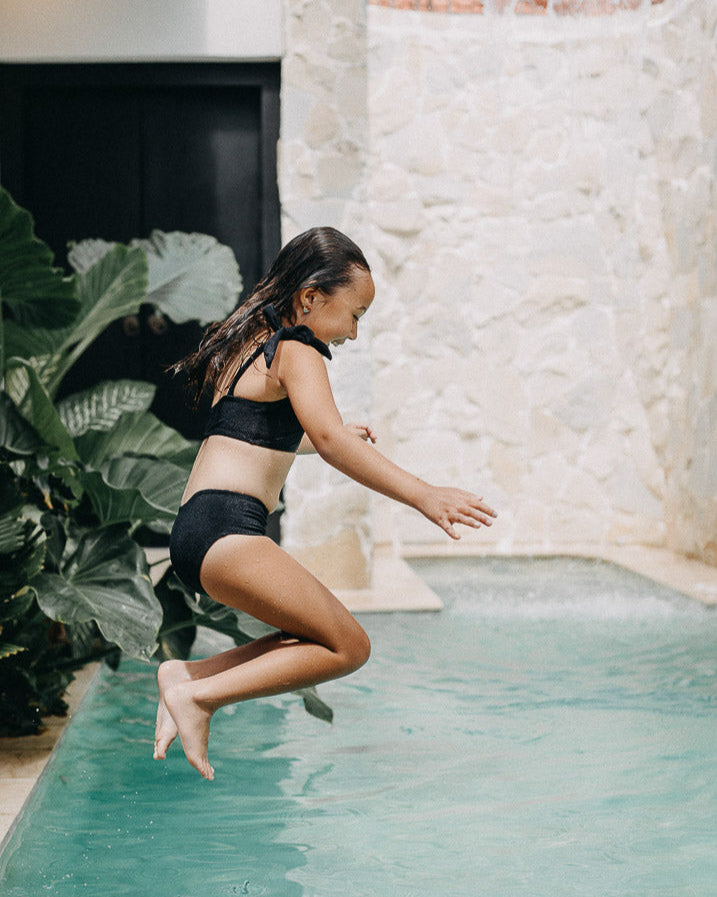 Ein junges Mädchen in einem Main Design Mia Mädchen Bikini springt mitten in einen türkisfarbenen Pool, umgeben von üppigen grünen Pflanzen und einer Steinmauer im Hintergrund. Sie scheint einen spielerischen Moment zu genießen, während Wasser in der Nähe plätschert. Der Badeanzug bietet LSF 50+-Schutz für sicheren Spaß unter der Sonne.