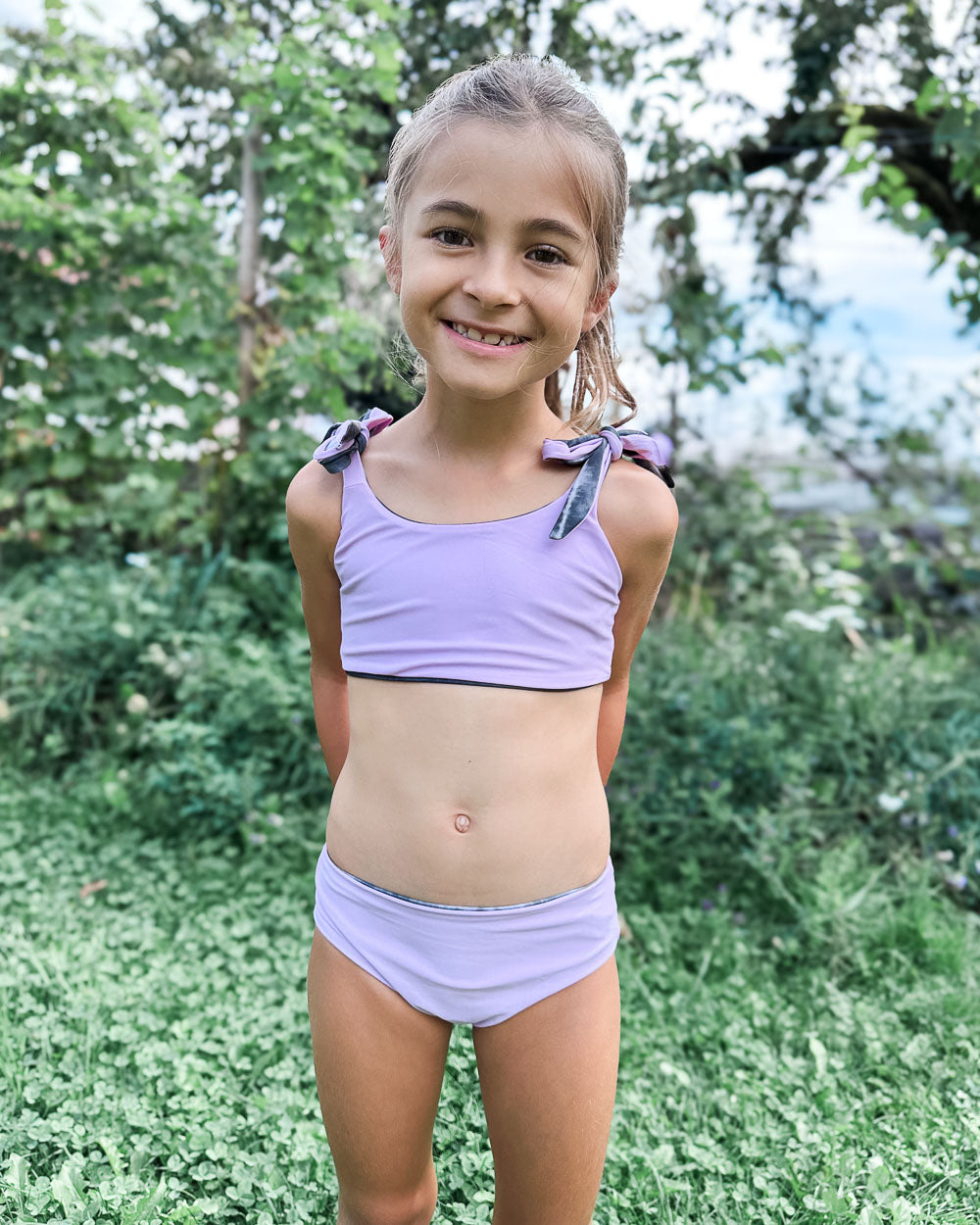 Ein junges Mädchen lächelt in die Kamera. Es steht auf grünem Gras und trägt den Mia Mädchen Bikini reversibel von Main Design - einen hellvioletten, nachhaltigen Bikini mit Schleifendetails. Im Hintergrund sind Bäume und Pflanzen zu sehen.