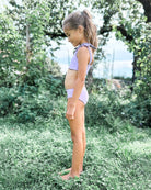 Ein Mädchen steht barfuß im Garten auf Gras und trägt den reversiblen, nachhaltigen Mia Mädchen Bikini in Helllila von Main Design. Sie ist im Profil zu sehen und blickt nach unten, umgeben von Bäumen und grünen Blättern.