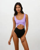 Eine Frau posiert vor einem schlichten weißen Hintergrund und trägt den Main Design Nova Badeanzug, einen ärmellosen einteiligen Badeanzug mit lavendelfarbenem Oberteil, schwarzem Unterteil und einem großen Ausschnitt an der Taille.