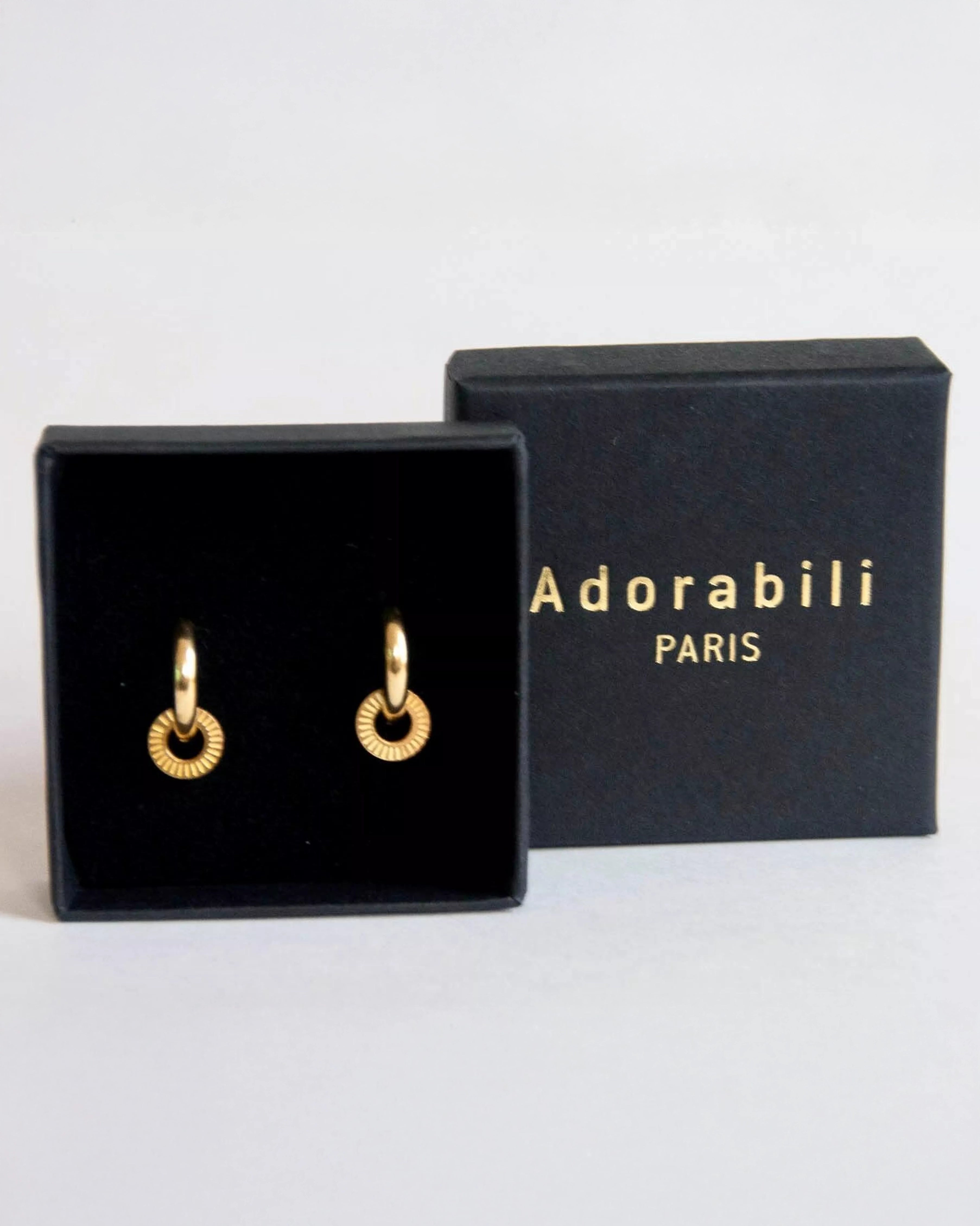 Ein Paar kleine Ohrringe Kreolen von Adorabili, 24 Karat vergoldet mit strukturierten Details, wird in einer schwarzen Schmuckschachtel neben einer passenden Adorabili Markenschachtel präsentiert.