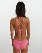 Eine Frau blickt von der Kamera abgewandt in dem Main Design Pasta Bikini Oberteil in Pink mit gekreuzten Trägern und einer Schleife hinten. Es ist aus chlorresistentem Stoff gefertigt und hat einen schlichten, hellen Hintergrund.