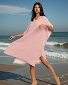 Eine Frau in einem lockeren, hellrosa Shoreline Breeze Poncho von Main Design läuft barfuß über einen Sandstrand mit sanften Wellen unter blauem Himmel.