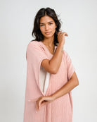 Eine Frau mit langen dunklen Haaren modelliert den lässigen Shoreline Breeze Poncho von Main Design in Hellrosa vor einem schlichten Hintergrund. Eine Größe passt den meisten, und sie posiert selbstbewusst mit einem erhobenen Arm.