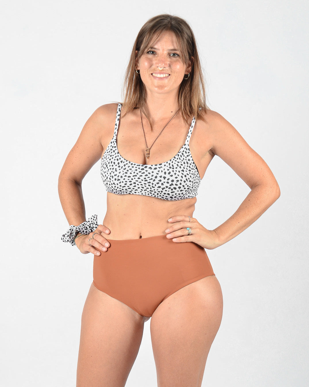 Eine Frau steht vor einem schlichten Hintergrund, lächelt und schaut in die Kamera. Sie trägt ein schwarz-weißes Bikinioberteil mit Punkten und Main Designs Posh Bikini Hose, eine hoch taillierte braune Bikinihose aus umweltfreundlichem Stoff. Sie hat ein Haargummi am Handgelenk und trägt Halsketten und Ringe für einen nahtlos stylischen Look.