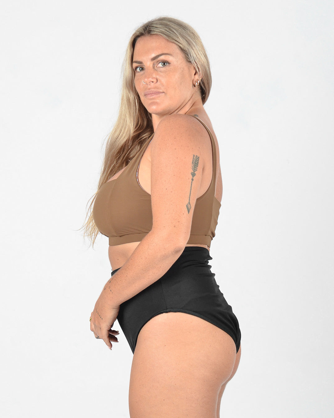 Eine Person mit langen blonden Haaren steht seitlich und blickt in die Kamera. Sie trägt einen braunen Sport-BH und eine hoch taillierte schwarze Hose aus umweltfreundlichem Stoff von Main Design namens Posh Bikini Hose. Der Hintergrund ist schlicht weiß.