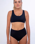 Vor einem schlichten weißen Hintergrund trägt eine Person mit zurückgebundenem langen Haar einen schwarzen Sport-BH aus recycelten Materialien, kombiniert mit der Posh Bikini Hose von Main Design – einem nahtlosen Unterteil mit hoher Taille. Sie zeigt kleine Tattoos auf jedem Arm und behält einen neutralen Ausdruck bei.