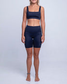 Eine Person steht barfuß vor einem schlichten Hintergrund und trägt einen schwarzen Sport-BH und schwarze Main Design Rena Bike Shorts aus recycelten Materialien. Sie hat schulterlanges blondes Haar, ein leichtes Lächeln und ihre Arme ruhen an den Seiten.