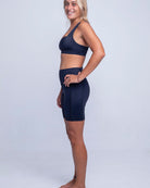 Eine Frau steht seitlich und blickt lächelnd in die Kamera. Sie trägt einen dunklen Sport-BH und dazu passende Rena Bike Shorts von Main Design aus recycelten Materialien. Sie steht barfuß vor einem schlichten, hellgrauen Hintergrund.