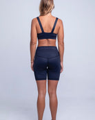 Eine Frau mit blonden Haaren steht mit dem Rücken zur Kamera. Sie trägt einen dunkelblauen Sport-BH und dazu passende hoch taillierte Rena Bike Shorts von Main Design aus recycelten Materialien. Der Hintergrund ist schlicht und neutral.