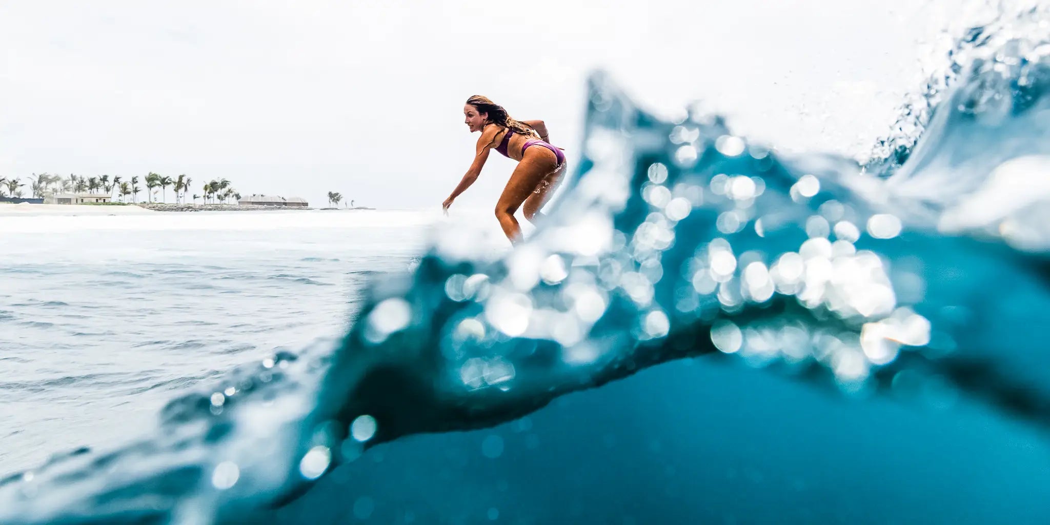 Eine Frau in einem lila Badeanzug surft auf einer Welle im Meer. Die Welle ist kristallklar und verdeckt den Surfer teilweise, wodurch eine dynamische und energiegeladene Szene entsteht. Der Hintergrund zeigt einen hellen Himmel und eine entfernte Küstenlinie mit Palmen und Gebäuden.