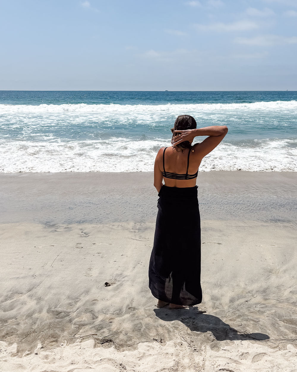Eine Frau in einem schwarzen Kleid und dem Sarong Bali von Main Design steht barfuß am Sandstrand und blickt mit geschlossenen Augen auf die Wellen unter einem teilweise bewölkten Himmel - ein perfekter Moment mit ihrem Lieblingsaccessoire.