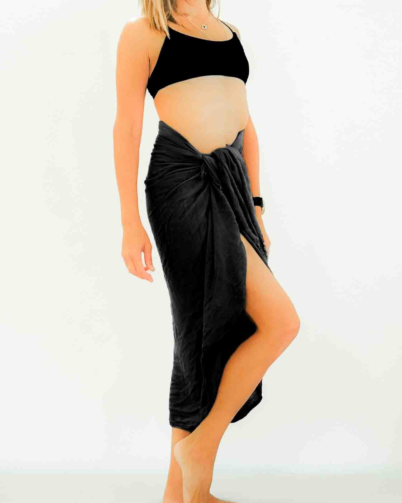 Mit dem Sarong Bali von Main Design präsentiert eine Person selbstbewusst ein schickes schwarzes bauchfreies Top, kombiniert mit einem dunklen Wickelrock, der an einen traditionellen Bali-Sarong erinnert, komplett mit einem stylischen Schlitz in der Taille vor einem schlichten Hintergrund.