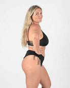 Eine Person mit langen blonden Haaren steht im Profil nach links und trägt das Sassy Bikini-Top von Main Design aus recycelten Materialien. Der schwarze Bikini betont ihre Figur mit seinem nahtlosen Schnitt vor einem schlichten weißen Hintergrund.