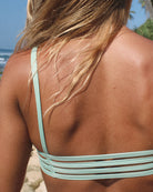 Am Strand trägt eine Person mit langen blonden Haaren das Sassy Bikini-Top von Main Design in Hellgrün, das aus recycelten Materialien hergestellt wurde. Sein nahtloses Design ergänzt perfekt das Meer und das Grün im Hintergrund.