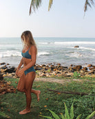 Eine Person im frechen Bikini-Top von Main Design steht auf einer Rasenfläche an einem sonnigen, tropischen Strand mit Wellen im Hintergrund und darüber hängenden Palmblättern.