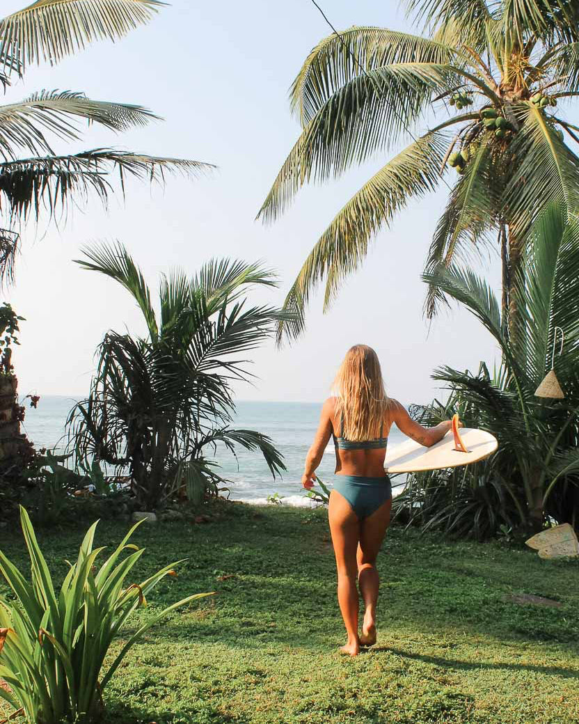 Eine Frau in einem Main Design Sassy Bikini-Top aus recycelten Materialien geht mit einem Surfbrett zum Meer. Üppiges Grün und Palmen umgeben sie unter einem klaren Himmel und schaffen eine tropische Atmosphäre.