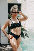 Eine Frau in einem Main Design Sassy Bikini-Top und Sonnenbrille steht lächelnd am Pool, die Haare hochgesteckt und die Sonnenbrille in der Hand. Sonnenlicht und grüne Pflanzen bilden einen lebendigen Hintergrund.