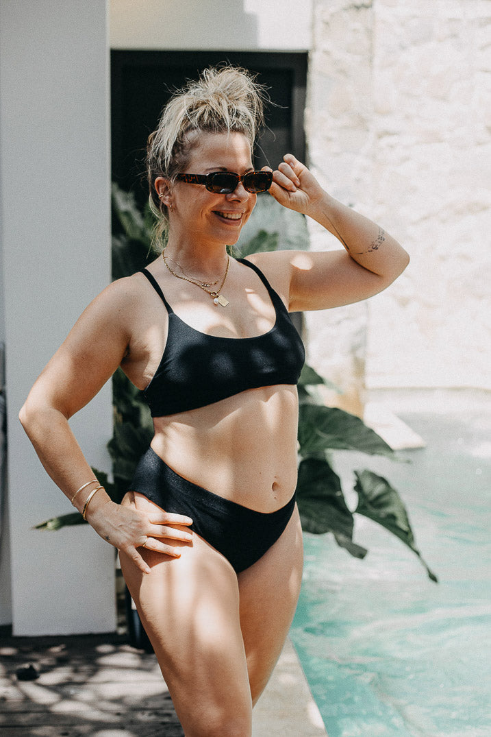 Eine Frau in einem Main Design Sassy Bikini-Top und Sonnenbrille steht lächelnd am Pool, die Haare hochgesteckt und die Sonnenbrille in der Hand. Sonnenlicht und grüne Pflanzen bilden einen lebendigen Hintergrund.