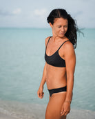 Eine Frau im Sassy Bikini-Top von Main Design steht an einem Sandstrand, ihr dunkles Haar ist vom Wind zerzaust, während sie nach unten blickt. Das Meer und der wolkenverhangene Himmel unterstreichen ihren sportlich-femininen Look aus recycelten Materialien.