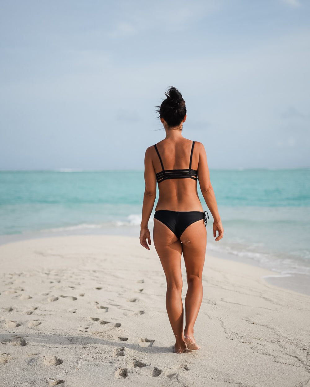 Eine Frau trägt das Sassy Bikini-Top von Main Design, das aus recycelten Materialien hergestellt wurde, während sie an einem Sandstrand steht und auf den türkisfarbenen Ozean unter einem meist klaren Himmel blickt, der eine entspannte tropische Stimmung erzeugt.