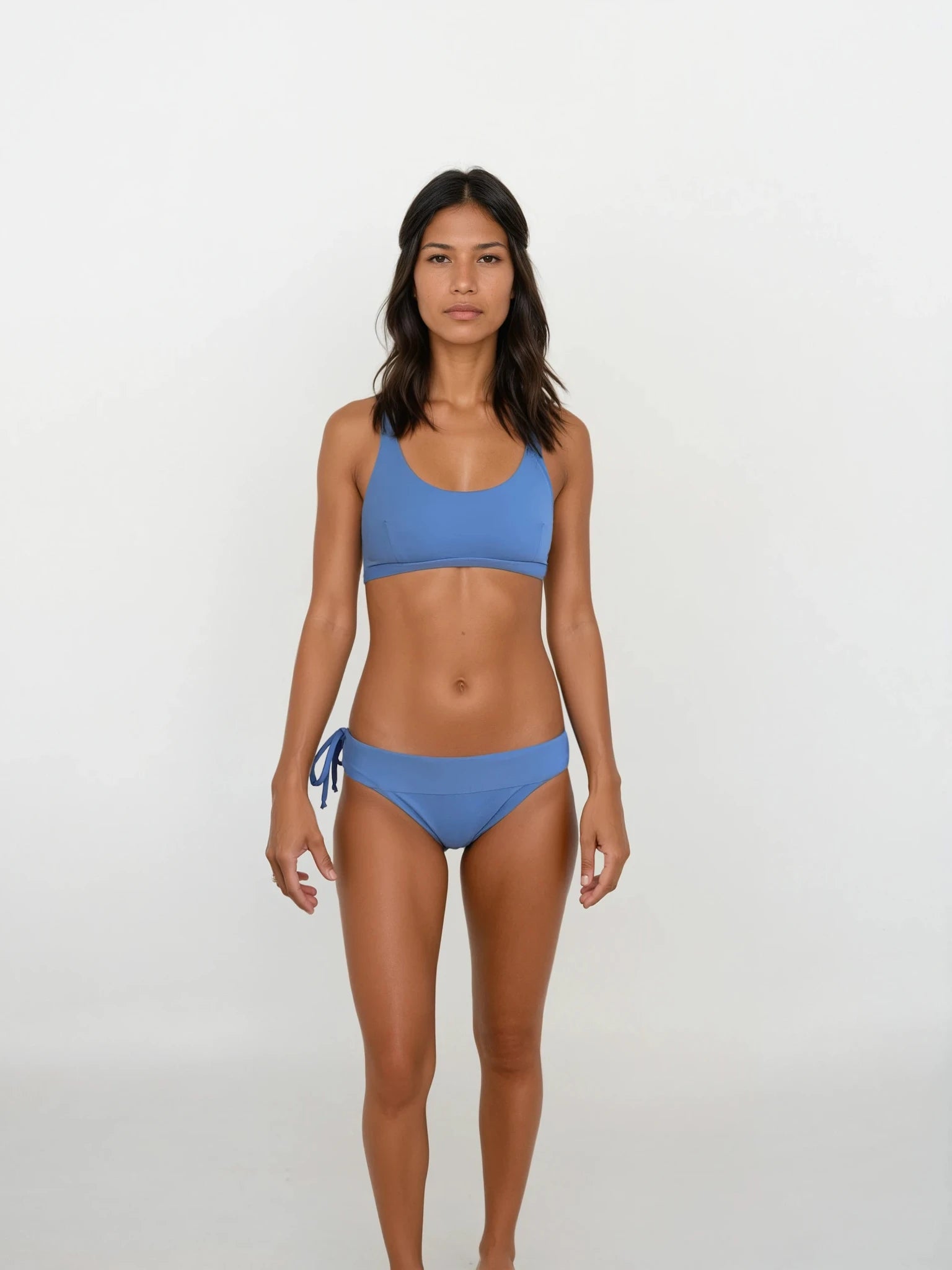 Eine Frau mit glattem, dunklem Haar und neutralem Gesichtsausdruck steht vor einem weißen Hintergrund. Sie trägt das Main Design Charm Surf Bikini Oberteil und eine blaue Hose mit seitlichen Bindebändern. Das Set bietet LSF 50+ Sonnenschutz – ideal für Wassersport.
