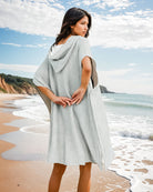 Eine Frau mit dem Shoreline Breeze Poncho von Main Design steht barfuß an einem Sandstrand und blickt auf das Meer. Klippen und Wellen bilden eine perfekte Kulisse für entspannte Strandtage unter einem teilweise bewölkten Himmel.