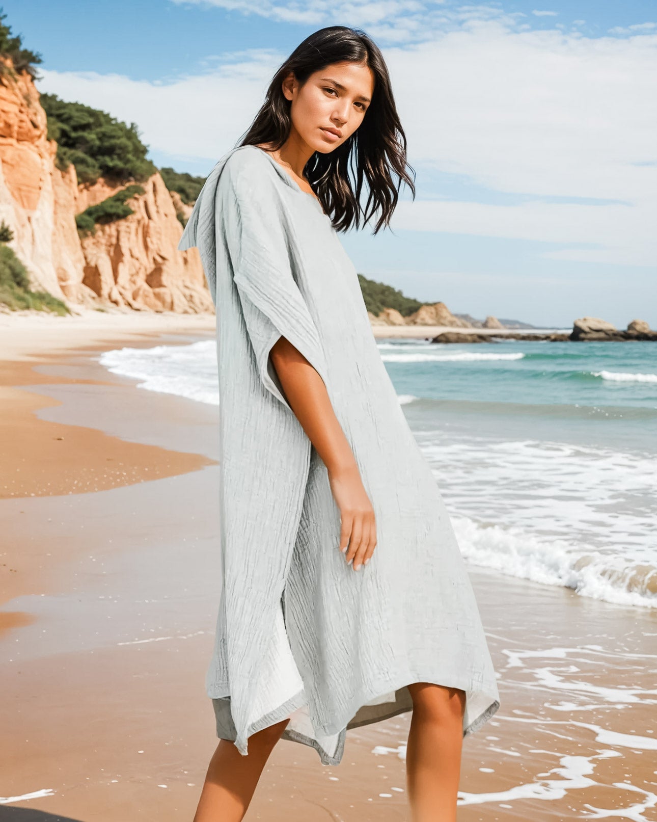 Eine Frau im Main Design Shoreline Breeze Poncho, einem lockeren hellgrauen Kleid, steht an einem Sandstrand mit Klippen und Meer im Hintergrund unter einem teilweise bewölkten Himmel - ideal für entspannte Strandtage.