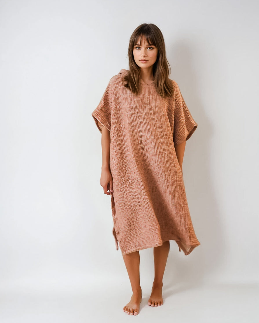 Eine Frau mit glattem braunem Haar steht barfuß vor einem schlichten weißen Hintergrund und trägt einen lockeren, strukturierten Shoreline Breeze Poncho in Hellbraun von Main Design. Sie blickt mit einem neutralen Blick nach vorne.