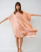 Eine Frau mit langen braunen Haaren steht barfuß, mit ausgestreckten Armen, und trägt einen lockeren, pfirsichfarbenen Shoreline Breeze Poncho von Main Design vor einem schlichten hellen Hintergrund.