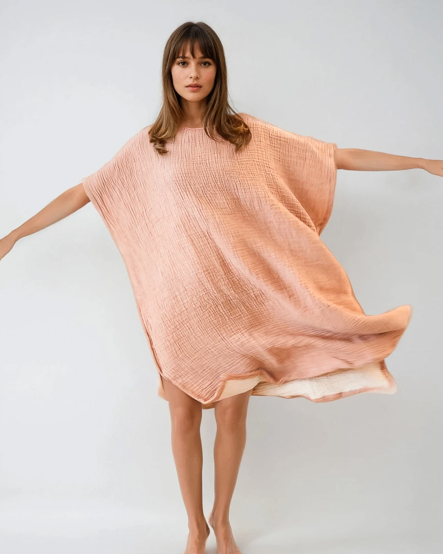 Eine Frau mit langen braunen Haaren steht barfuß, mit ausgestreckten Armen, und trägt einen lockeren, pfirsichfarbenen Shoreline Breeze Poncho von Main Design vor einem schlichten hellen Hintergrund.