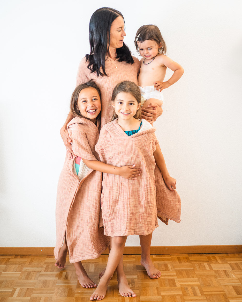 Eine Frau lächelt mit drei kleinen Kindern. Zwei Mädchen tragen zusammenpassende rosa Main Design Shoreline Breeze Ponchos, während ein Kleinkind in einer Windel in ihren Armen gehalten wird. Alle stehen barfuß auf einem Holzboden vor einer weißen Wand.