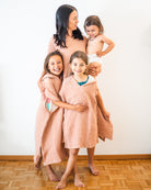 Eine Frau lächelt mit drei kleinen Kindern. Zwei Mädchen tragen zusammenpassende rosa Main Design Shoreline Breeze Ponchos, während ein Kleinkind in einer Windel in ihren Armen gehalten wird. Alle stehen barfuß auf einem Holzboden vor einer weißen Wand.