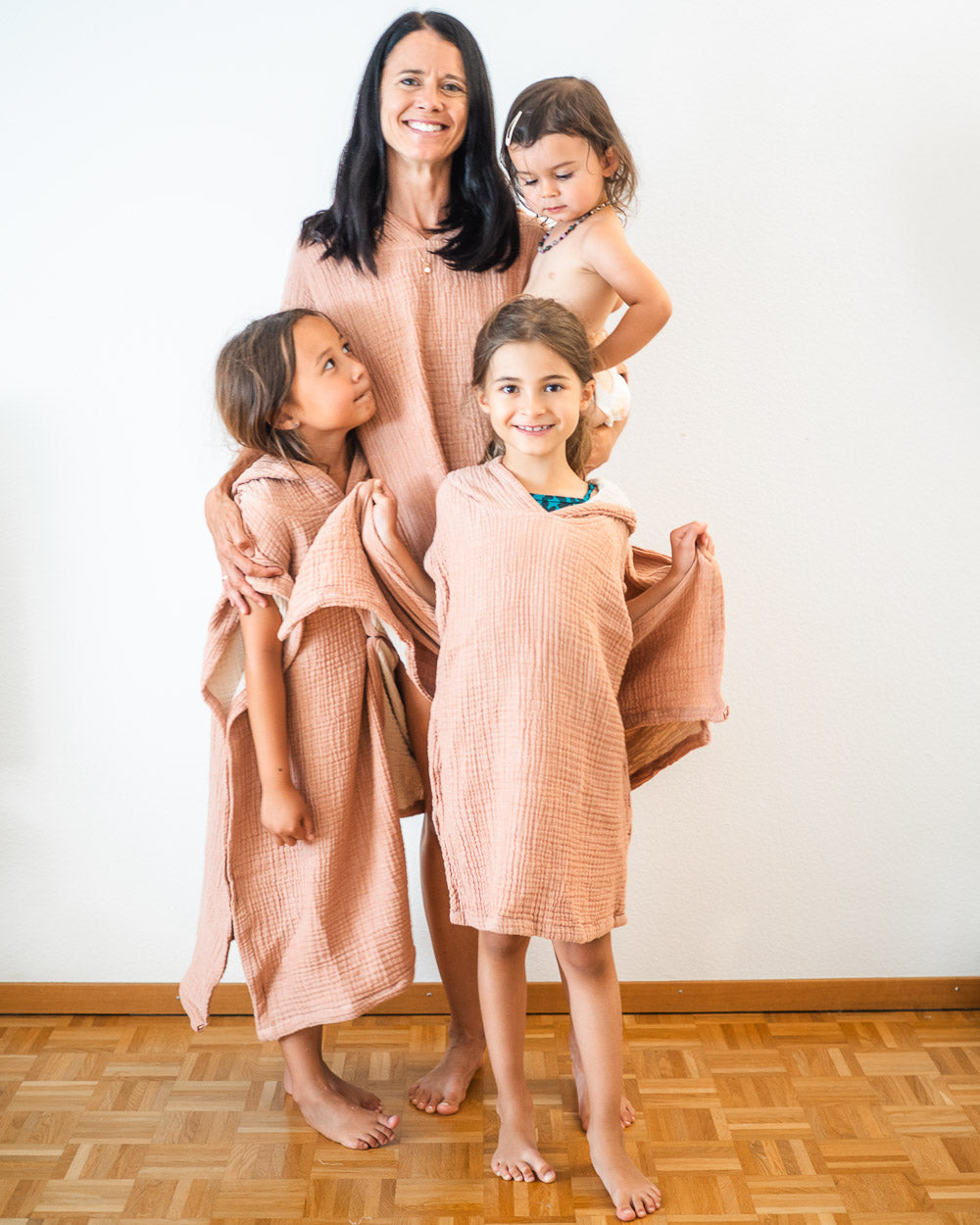 Eine Frau lächelt mit drei barfüßigen Kindern, die alle passende Main Design Shoreline Breeze Poncho-Kleidung tragen. Ihre kuscheligen Einheitsgrößen passen zu dem einfachen Holzboden und der schlichten weißen Wand hinter ihnen.