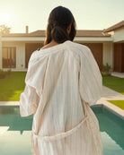 Eine Frau mit dunklem Haar steht an einem modernen Außenpool, von hinten gesehen, und trägt den hellen Soleil Kimono von Main Design. Die sonnenbeschienene Umgebung unterstreicht den grünen Rasen des Hauses und die nachhaltigen Produktionswerte.