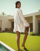 Eine Frau in einem lockeren, hellen Soleil-Kimono von Main Design aus 100 % Baumwolle steht barfuß im Gras in der Nähe eines Pools und blickt über ihre Schulter. Im Hintergrund erheben sich moderne Gebäude und Palmen unter einem klaren Himmel.