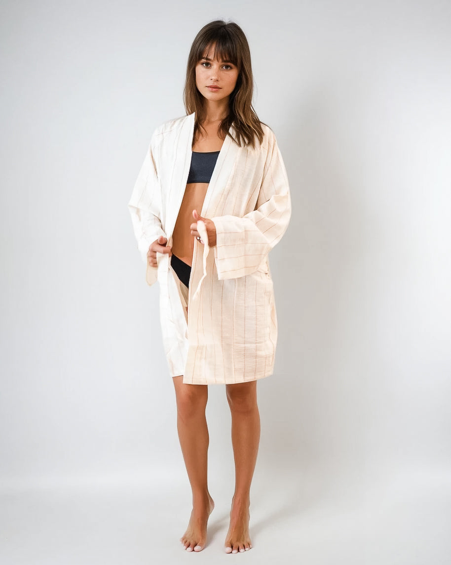 Eine Frau steht barfuß vor einem schlichten Hintergrund, trägt den Main Design Soleil Kimono - einen hellen, lockeren Bademantel aus 100 % Baumwolle - über einem schwarzen Bikini oder Unterwäsche und blickt mit neutralem Blick direkt in die Kamera.