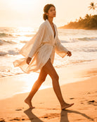 Eine Frau in dem leichten, fließenden Soleil Kimono von Main Design aus nachhaltiger Baumwolle spaziert barfuß am Strand bei Sonnenuntergang mit Wellen, Palmen und goldenem Sonnenlicht im Hintergrund.